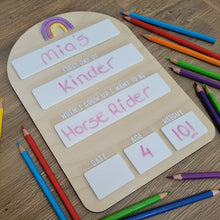 Load image into Gallery viewer, A wooden first day of school board with slots for labels, currently filled with 'Mia's First Day of Kinder', 'Horse Rider', and other placeholders. The board is arched at the top and has a rainbow and sun graphic at the top left corner.