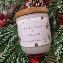 Load image into Gallery viewer, Candle labeled 'Frankincense &amp; Myrrh' with a wooden lid, surrounded by festive greenery and red berries.
