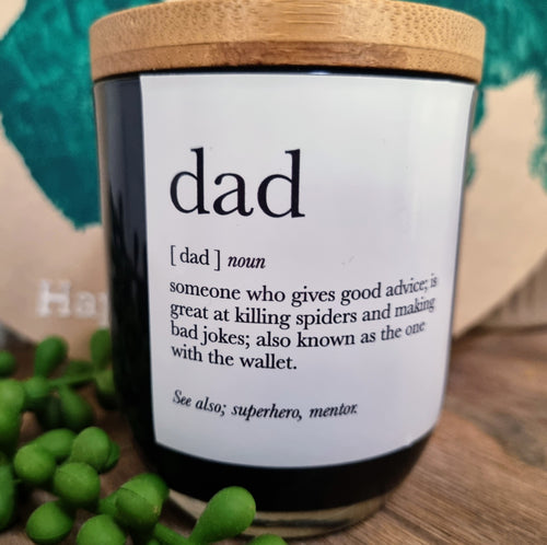 A black candle with a wooden lid, labeled 'DAD' with a definition below it, placed on a wooden surface with greenery in the background.