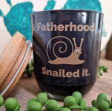 Load image into Gallery viewer, A black soy candle with a lid, featuring a dad joke text and a graphic of a snail with the phrase &#39;Fatherhood Snailed it.&#39;
