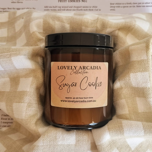 Candle jar labeled 'Sugar Cookie' from the 'Lovely Arcadia' collection on a textured fabric background.