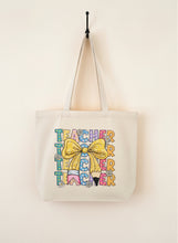 Load image into Gallery viewer, Beige tote bag with colorful &#39;Teacher&#39; text and a yellow bow on a wooden surface.
