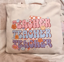 Load image into Gallery viewer, Beige tote bag with colorful &#39;Teacher Teacher Teacher&#39; text and graphics on a soft surface.
