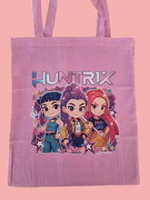 Load image into Gallery viewer, Pink tote bag with cartoon characters and &#39;HUNTRIX&#39; branding on a beige surface

